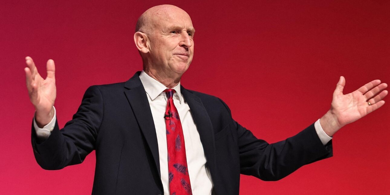 Chancellor John Healey - a white man wearing a dark suit - stood against a red background with his arms open wide.